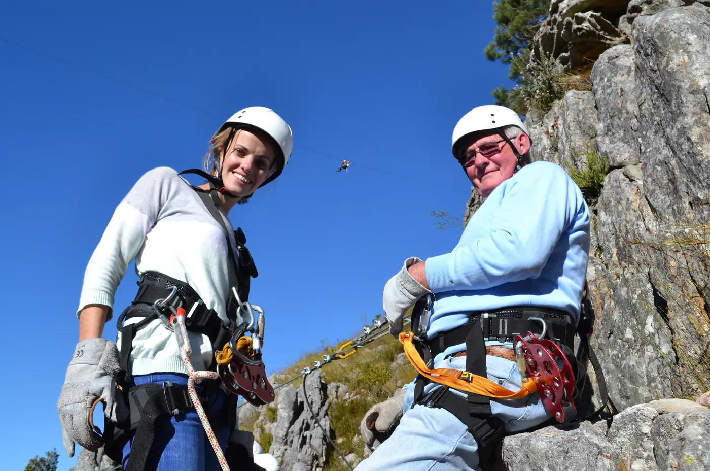 A grandfather and his granddaughter in helmets and harnesses on a Cape Canopy Tour zipline platform, with another zipliner crossing the valley behind them